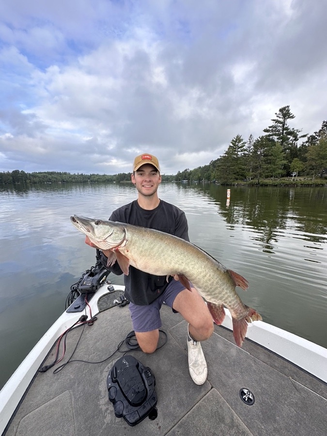 Phillip kneeling on the bow of a boat on a calm lake, holding a large muskie